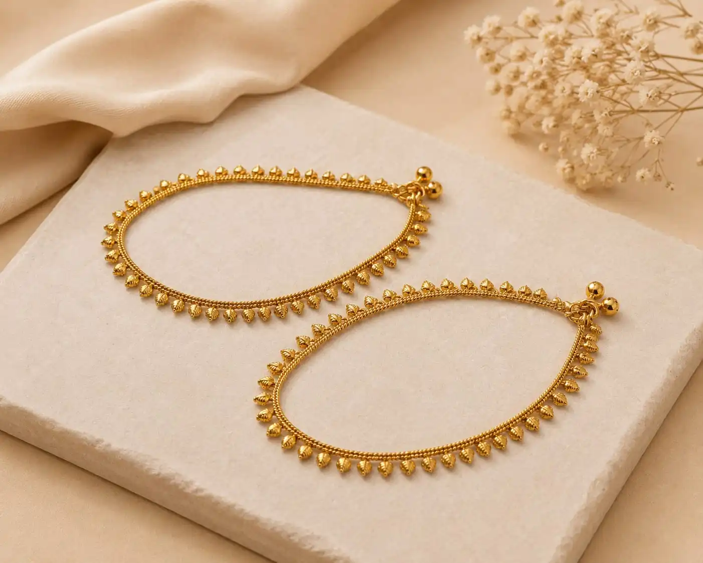Golden Bead Grace Payal featuring delicate gold-tone chain detailing and evenly spaced teardrop-shaped bead accents, displayed on a cream stone surface with soft beige fabric and dried flowers.
