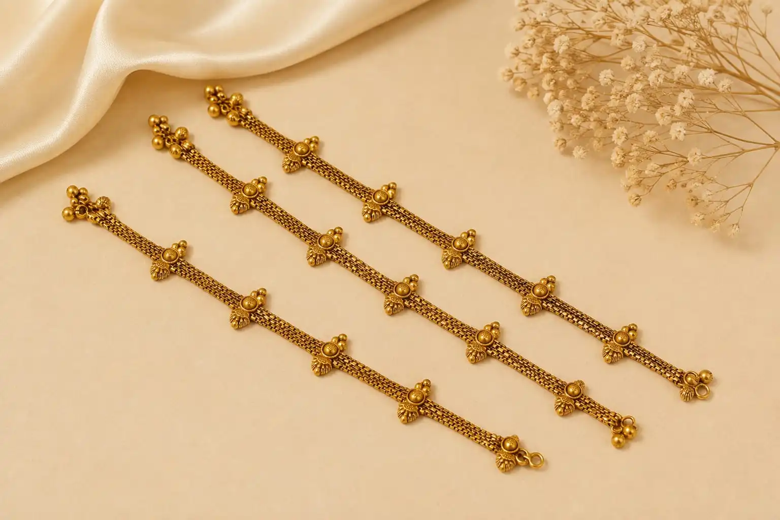 Royal Floral Beaded Gold Tone Payal featuring delicate chainwork, repeated floral motifs and small ornamental bead details, displayed on a soft beige background with satin fabric and dried flowers.