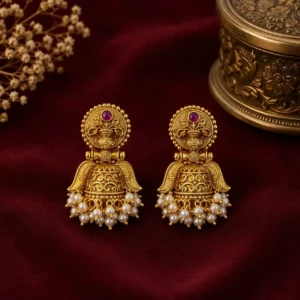 Pair of traditional gold-tone jhumka earrings with intricate floral detailing, ruby-toned accents, and pearl-style bead fringes displayed on rich burgundy fabric.