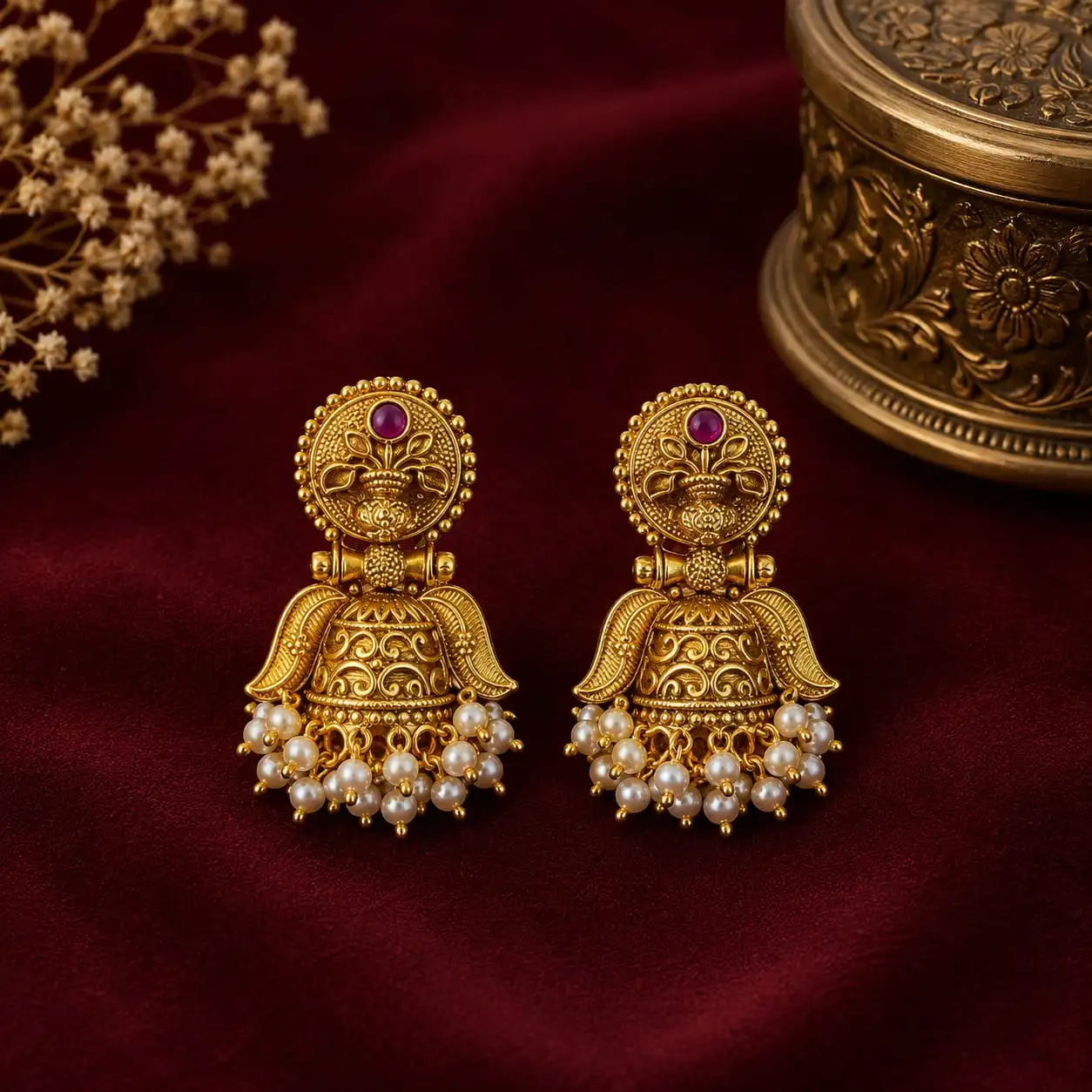 Pair of traditional gold-tone jhumka earrings with intricate floral detailing, ruby-toned accents, and pearl-style bead fringes displayed on rich burgundy fabric.