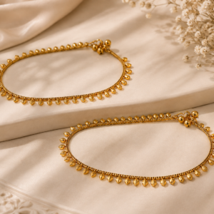 Gold-tone traditional payal with delicate chainwork and evenly spaced leaf-shaped dangling charms, displayed on soft cream fabric with a minimal elegant decorative background and dried flowers.