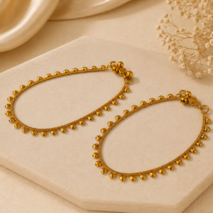 Golden Bead Grace Payal featuring delicate gold-tone chainwork and repeated teardrop-shaped bead charms, displayed on a cream stone platform with soft beige satin fabric and dried flowers.
