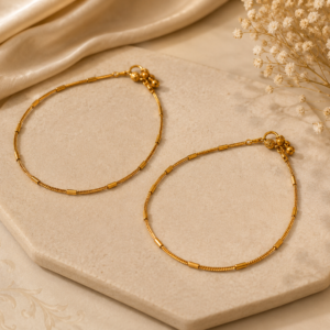 Gold-tone payal featuring a delicate chain with evenly spaced cylindrical tube-style details and small decorative clasp charms, showcased on a beige stone platform with soft satin fabric and dried flowers.