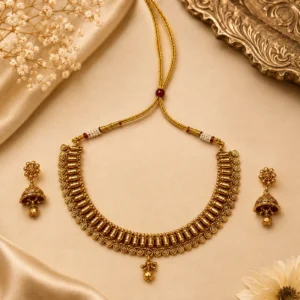 Traditional gold-tone necklace set with matching jhumka earrings, featuring intricate circular motifs, layered bead detailing, red and white accents, and a decorative pendant displayed on elegant beige satin with flowers.