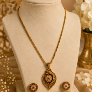 "Traditional gold-tone necklace with ornate teardrop pendant and matching round stud earrings displayed on a cream jewelry "