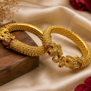 Elegant gold-tone traditional bangles with intricate detailing and red stone accents displayed on cream satin.