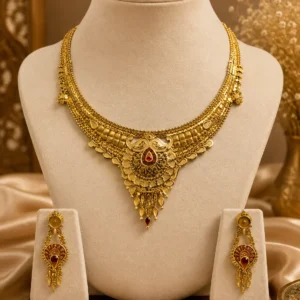 Gold-tone traditional necklace set displayed on a cream jewellery mannequin, featuring intricate layered detailing, a central red teardrop stone pendant, and matching long drop earrings on a warm beige background.