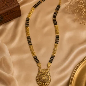 Gold-tone black beaded traditional necklace displayed on champagne-colored satin, featuring intricate ornamental detailing, decorative gold panels, and an ornate central pendant with layered hanging elements.