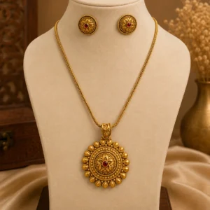 Gold-tone floral pendant necklace set displayed on a cream jewellery mannequin, featuring a large circular pendant with intricate floral detailing and red centre stone, paired with matching round stud earrings against a warm beige background.