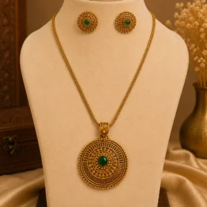 Gold-tone traditional necklace set displayed on a cream jewellery mannequin, featuring a large circular pendant with intricate floral detailing and a green centre stone, paired with matching round green-stone earrings against a warm beige background.