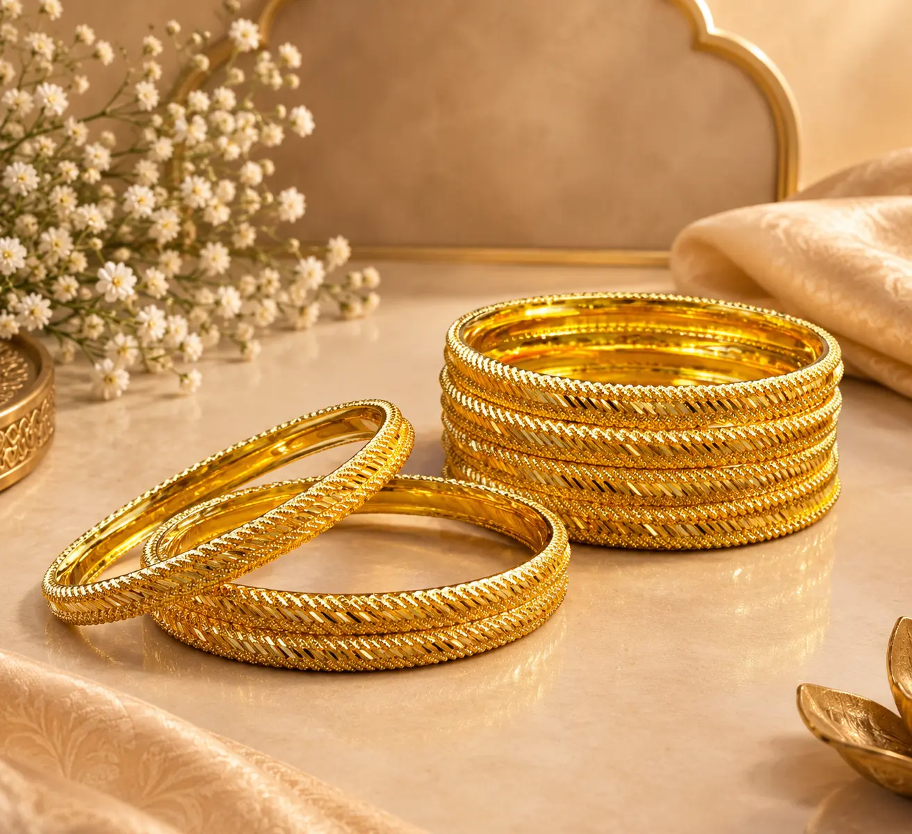 Gold-tone traditional bangles with intricate braided and beaded detailing displayed on a luxurious beige surface with silk fabric and floral accents.