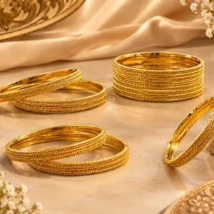 Gold-tone traditional bangles with intricate braided, beaded, and textured detailing arranged on a luxurious beige surface with soft satin fabric and white floral accents.