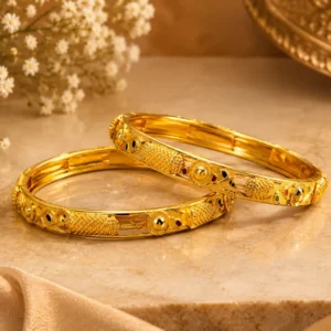 Two elegant gold-tone bangles with intricate textured, beaded and engraved detailing displayed on a luxurious beige marble surface with soft floral and satin accents.