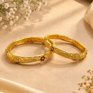 Two elegant gold-tone traditional bangles with intricate floral, engraved, and textured detailing displayed on a luxurious beige surface with soft satin fabric and white floral accents.