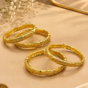 Four gold-tone traditional bangles with intricate geometric, engraved, and textured detailing arranged elegantly on a luxurious beige surface with soft satin fabric and white floral accents.