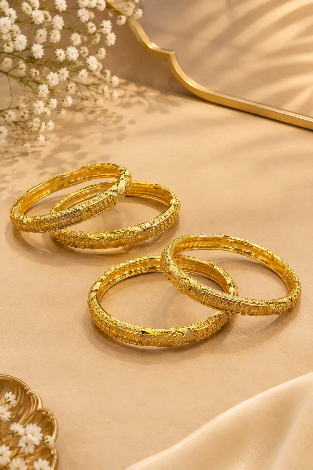 Four gold-tone traditional bangles with intricate geometric, engraved, and textured detailing arranged elegantly on a luxurious beige surface with soft satin fabric and white floral accents.