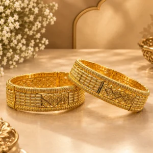 Two gold-tone traditional bangles with intricate geometric lattice patterns, beaded borders, engraved detailing, and raised ornamental accents displayed elegantly on a luxurious beige surface with soft floral and golden décor.