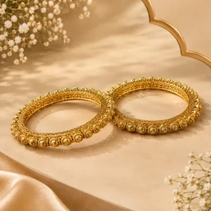 Gold-tone ornate bangles with intricate floral detailing displayed on a beige surface with soft satin and white floral accents.