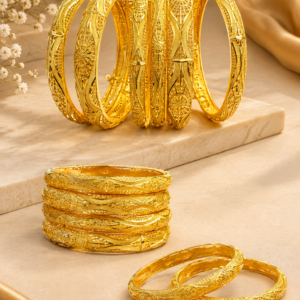 Gold-tone decorative bangles with intricate floral, geometric and engraved patterns arranged on a cream textured display surface, featuring stacked and overlapping designs in a warm luxury jewellery showcase.