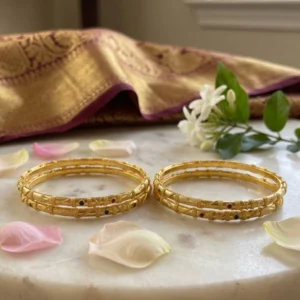 Two pairs of traditional gold bangles featuring delicate patterned texturing and embedded colored stones, elegantly displayed on a white marble slab surface next to scattered flower petals and a draped silk fabric.
