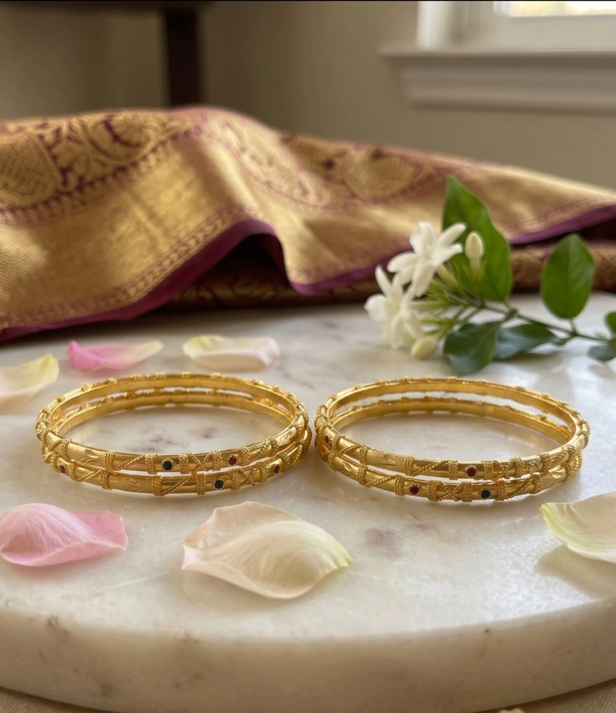 Two pairs of traditional gold bangles featuring delicate patterned texturing and embedded colored stones, elegantly displayed on a white marble slab surface next to scattered flower petals and a draped silk fabric.